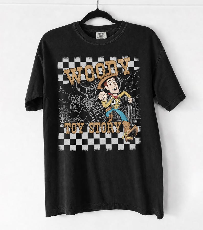 May include: A black short-sleeve t-shirt featuring a graphic print of the character Woody from Toy Story. The design includes a black and white checkered border, a desert background, and the text WOODY TOY STORY in a tan, distressed font style.
