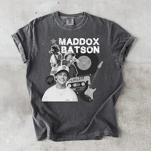 May include: Dark gray t-shirt featuring a black and white graphic of a person, a record, a guitar, and the text "MADDOX BATSON" and "PLAYLIST". The shirt has a crew neck and short sleeves.