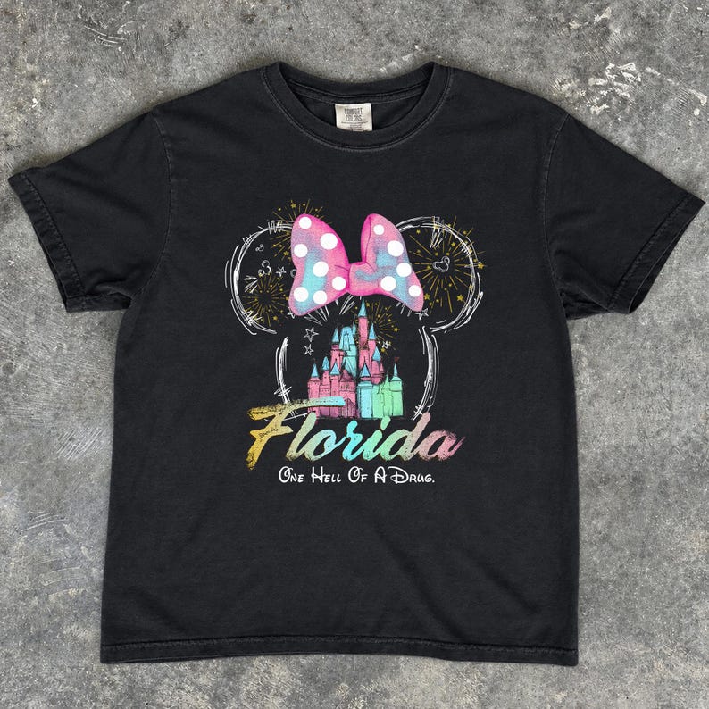 May include: Black t-shirt featuring a colorful graphic of a castle, fireworks, and a polka-dotted bow in the shape of Mickey Mouse ears. The word "Florida" is written in a rainbow font, with the phrase "One Hell Of A Drug" below.