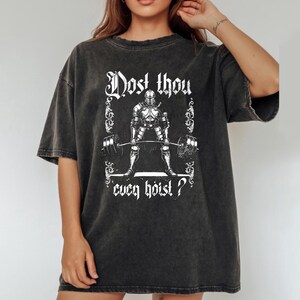 May include: Dark grey t-shirt featuring a white graphic of a knight lifting a barbell. The text "Dost thou even hoist?" is printed above and below the knight illustration. The shirt has a vintage, washed appearance.
