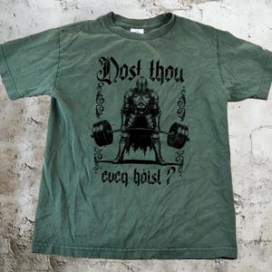 May include: Green t-shirt featuring the phrase "Dost thou even hoist?" and a black graphic of a knight lifting a barbell. The shirt has a crew neck and a vintage look.
