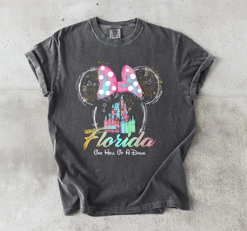 May include: Dark gray t-shirt featuring a Minnie Mouse design with a polka dot bow, fireworks, and a castle. The word "Florida" is written in a colorful font, with the phrase "One Hell Of A Drug" below.