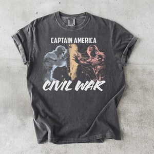 May include: Dark gray t-shirt featuring a graphic from Captain America: Civil War. The design shows two figures in action, with the words "CAPTAIN AMERICA" above and "CIVIL WAR" below in white. The shirt has a vintage, worn look.
