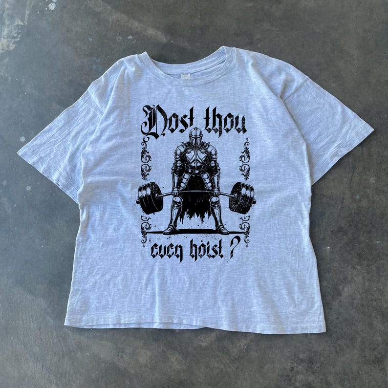 May include: Pale grey t-shirt with a black illustration of a knight lifting a barbell. The text "Dost thou ever hoist?" is above and below the knight graphic. The shirt is made of a soft material.