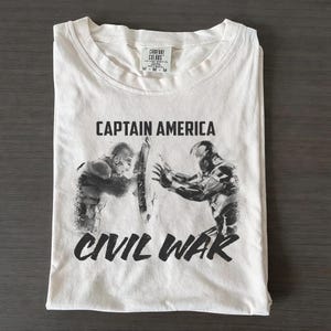 May include: White t-shirt featuring a graphic of Captain America and Iron Man from Civil War. The text "CAPTAIN AMERICA" is printed above the image, and "CIVIL WAR" is printed below. The shirt is folded neatly on a dark surface.