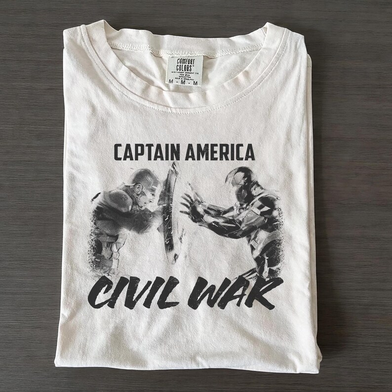 May include: White t-shirt featuring a graphic of Captain America and Iron Man from Civil War. The text "CAPTAIN AMERICA" is printed above the image, and "CIVIL WAR" is printed below. The shirt is folded neatly on a dark surface.