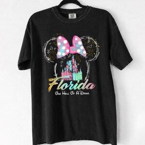 May include: Black t-shirt featuring a colorful graphic of a castle, a polka-dot bow, and the word "Florida" in a rainbow gradient. The text "One Hell Of A Drug" is below the word Florida.