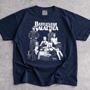 May include: Navy blue t-shirt with a graphic of the cast from Battlestar Galactica. The white text "Battlestar Galactica" is above the image of the cast. The t-shirt is made of a soft material.