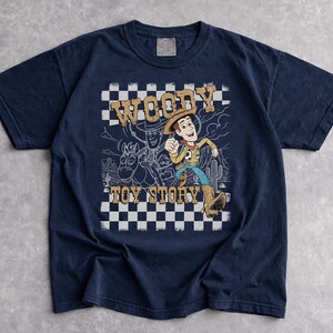 May include: A navy blue short-sleeve t-shirt featuring a graphic print of Woody from Toy Story. The design includes a black and white checkered background, a desert landscape outline, and the text WOODY TOY STORY in a tan, distressed font.