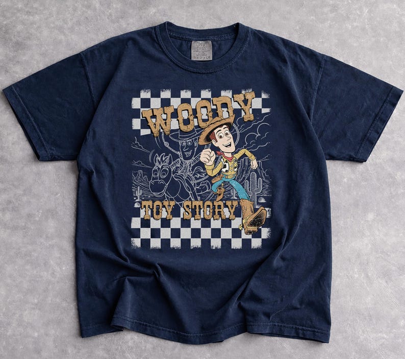 May include: A navy blue short-sleeve t-shirt featuring a graphic print of Woody from Toy Story. The design includes a black and white checkered background, a desert landscape outline, and the text WOODY TOY STORY in a tan, distressed font.
