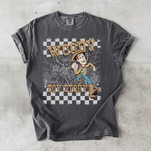 May include: A charcoal gray short-sleeve t-shirt featuring a graphic print of the character Woody from Toy Story. The design includes a black and white checkered background, a desert landscape outline, and the text Woody Toy Story in tan.