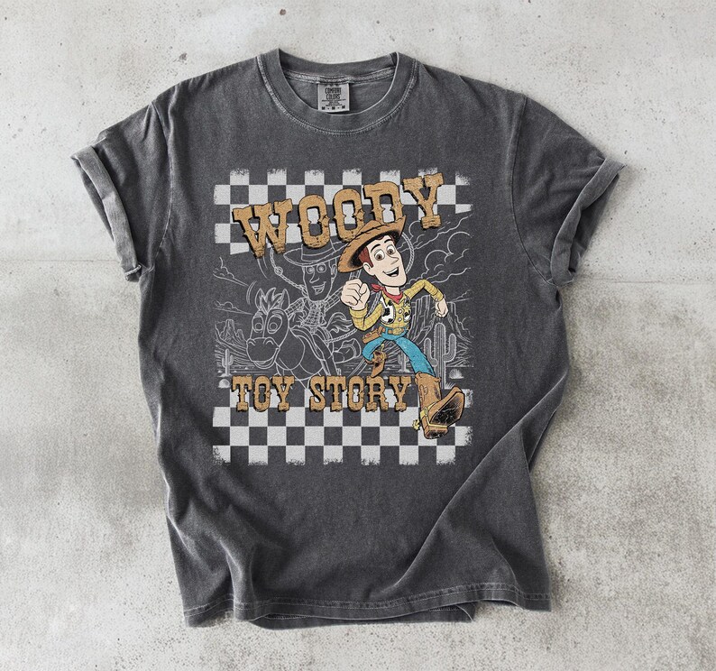 May include: A charcoal gray short-sleeve t-shirt featuring a graphic print of the character Woody from Toy Story. The design includes a black and white checkered background, a desert landscape outline, and the text Woody Toy Story in tan.