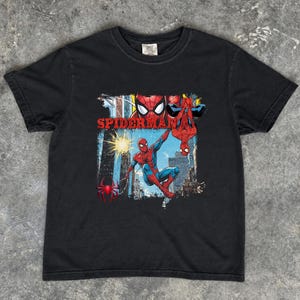 Puede incluir: Camiseta negra con un gr&aacute;fico colorido de Spider-Man. El dise&ntilde;o muestra a Spider-Man en poses de acci&oacute;n, con la palabra "SPIDERMAN" en rojo. El fondo representa un paisaje urbano. La camiseta tiene cuello redondo.