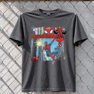 Puede incluir: Camiseta gris oscuro con un dise&ntilde;o de Spider-Man. El dise&ntilde;o muestra a Spider-Man en acci&oacute;n, con la palabra "SPIDERMAN" en rojo. El fondo muestra una escena urbana. El superh&eacute;roe lleva su traje rojo y azul.