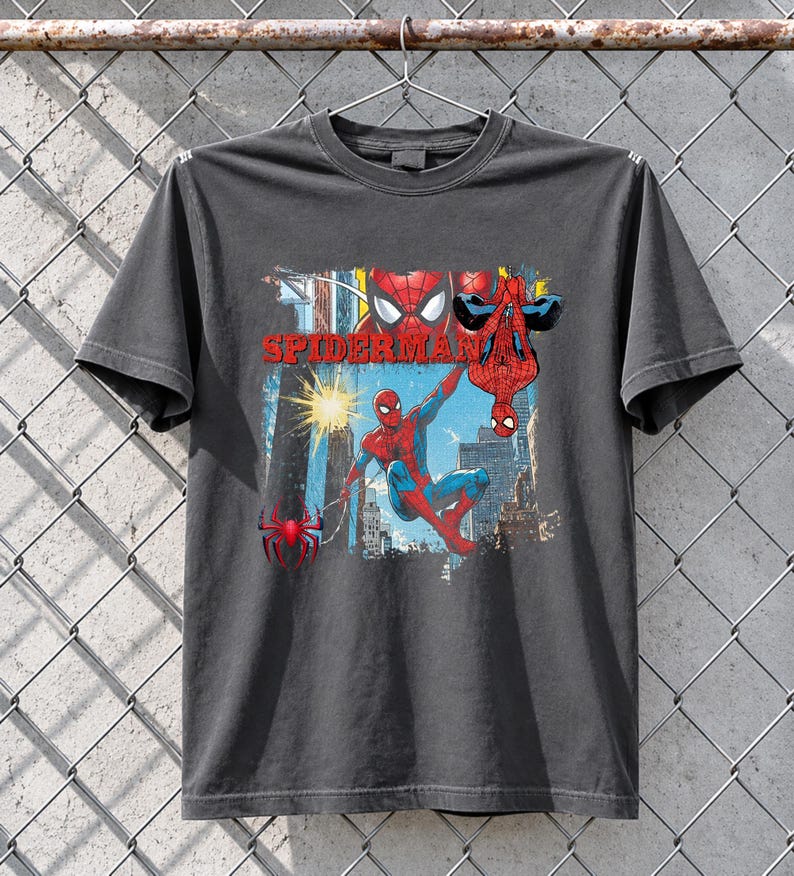 Puede incluir: Camiseta gris oscuro con un dise&ntilde;o de Spider-Man. El dise&ntilde;o muestra a Spider-Man en acci&oacute;n, con la palabra "SPIDERMAN" en rojo. El fondo muestra una escena urbana. El superh&eacute;roe lleva su traje rojo y azul.