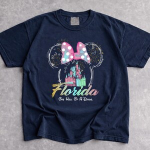 May include: Navy blue t-shirt featuring a Minnie Mouse design with a pink polka dot bow, a castle, and the word "Florida". The text "One Hell Of A Drug" is below the word Florida. The design is in white, pink, blue, and green.
