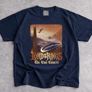 May include: Navy blue t-shirt featuring a graphic from "The Lord of the Rings: The Two Towers." The design includes a tower, a dragon, and the inscription "Lord of the Rings" and "The Two Towers" in a stylized font. The shirt is made of a soft material.