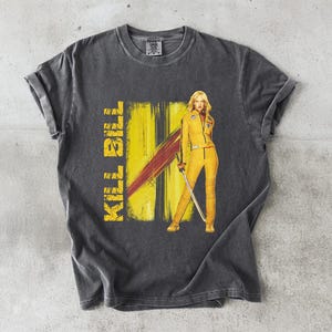 Op de afbeelding: Donkergrijs T-shirt met een afbeelding uit de film "Kill Bill". Het ontwerp bevat de tekst "KILL BILL" in geel, een gele en rode achtergrond en een vrouw in een gele overall met een zwaard.