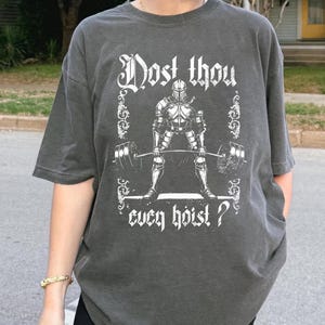 May include: Dark grey t-shirt featuring a knight lifting weights graphic, framed with decorative elements. The text reads "Dost thou even hoist?" in a stylized font. The shirt has short sleeves and a relaxed fit.