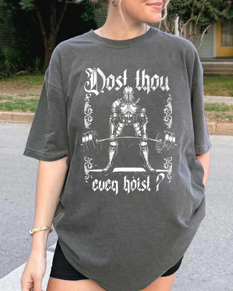May include: Dark grey t-shirt featuring a knight lifting weights graphic, framed with decorative elements. The text reads "Dost thou even hoist?" in a stylized font. The shirt has short sleeves and a relaxed fit.
