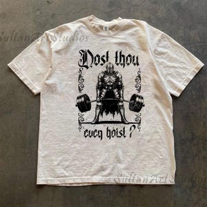 May include: Cream-coloured t-shirt with a black graphic of a knight lifting a barbell. The text "Dost thou ever hoist?" is above and below the knight. The shirt has short sleeves and a crew neck.