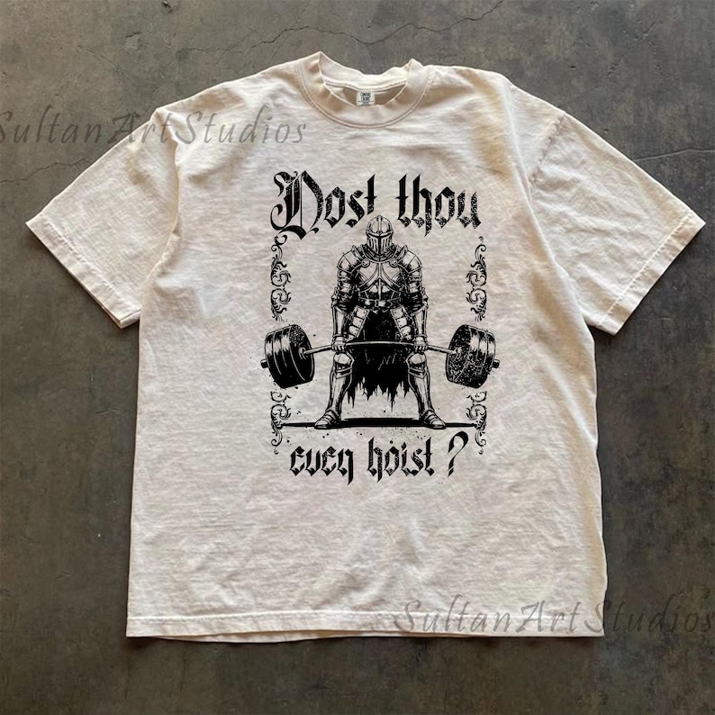 May include: Cream-coloured t-shirt with a black graphic of a knight lifting a barbell. The text "Dost thou ever hoist?" is above and below the knight. The shirt has short sleeves and a crew neck.