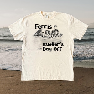 Könnte beinhalten: Cremefarbenes T-Shirt mit einer Schwarz-Weiß-Grafik eines Autos mit Personen darin und dem Text "Ferris Bueller's Day Off". Das T-Shirt wird an einem Strand mit dem Meer im Hintergrund präsentiert.