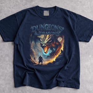 May include: Navy blue t-shirt featuring a fantasy illustration of a dragon and two figures with the text "DUNGEONS & DRAGONS". The graphic is in shades of orange, blue, and white. The shirt is made of a soft material.