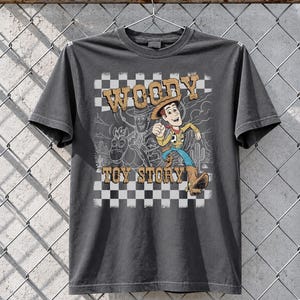 May include: A charcoal gray short-sleeve t-shirt featuring a graphic print of the character Woody from Toy Story. The design includes a checkered background, a desert landscape outline, and the text WOODY TOY STORY in a distressed brown font.