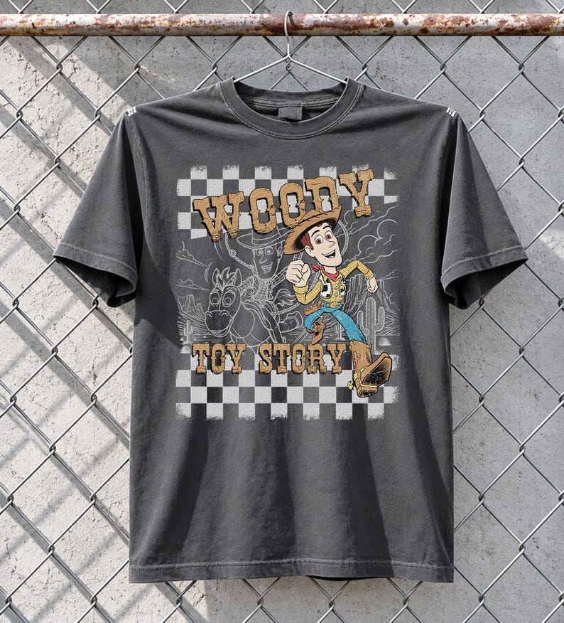 May include: A charcoal gray short-sleeve t-shirt featuring a graphic print of the character Woody from Toy Story. The design includes a checkered background, a desert landscape outline, and the text WOODY TOY STORY in a distressed brown font.