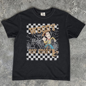 May include: A black short-sleeve t-shirt featuring a graphic print of the character Woody from Toy Story. The design includes a black and white checkered background, a desert landscape outline, and the text Woody Toy Story in tan lettering.