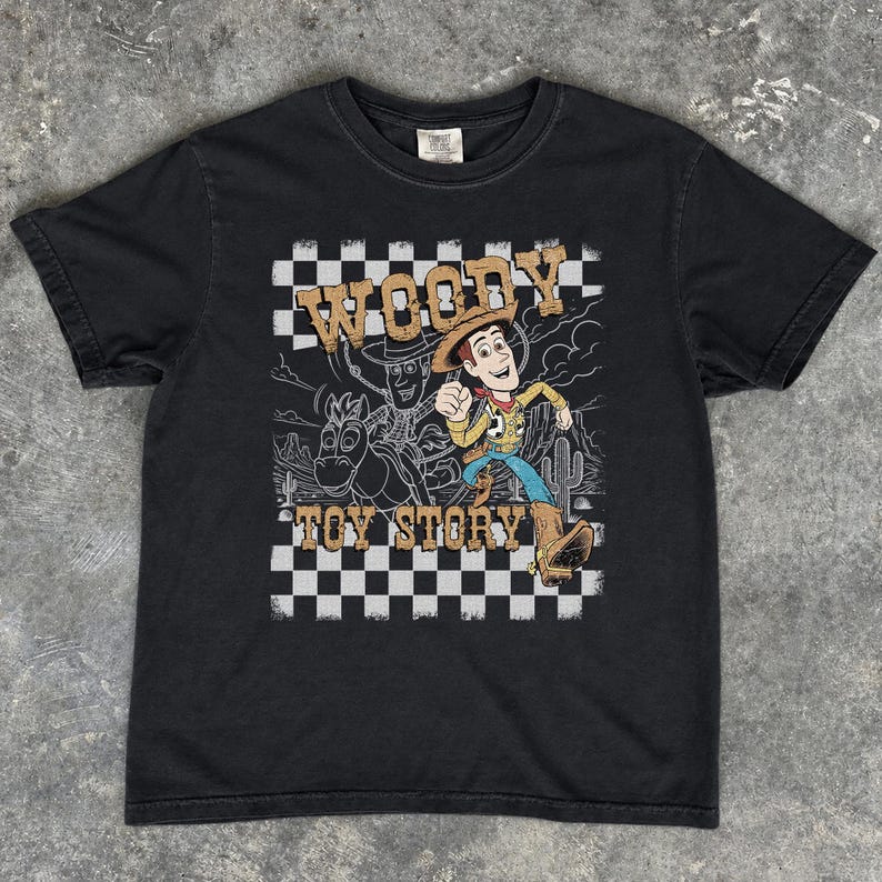 May include: A black short-sleeve t-shirt featuring a graphic print of the character Woody from Toy Story. The design includes a black and white checkered background, a desert landscape outline, and the text Woody Toy Story in tan lettering.