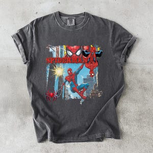 Puede incluir: Camiseta gris oscuro con un gr&aacute;fico colorido de Spider-Man. El dise&ntilde;o presenta a Spider-Man en rojo y azul, con la palabra "SPIDERMAN" en rojo. La camiseta tiene un aspecto vintage y desgastado, y est&aacute; hecha de un material suave.