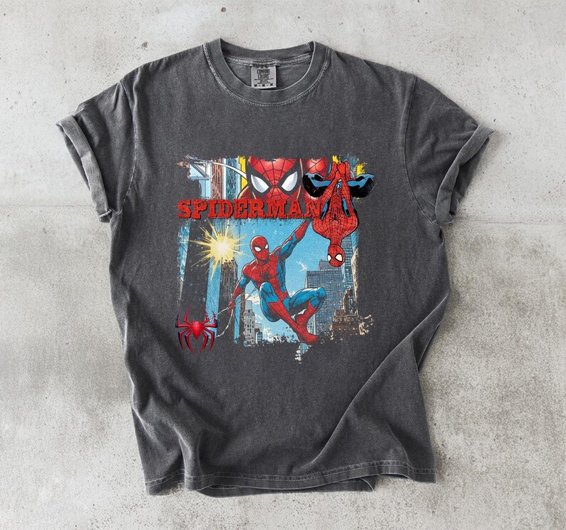 Puede incluir: Camiseta gris oscuro con un gr&aacute;fico colorido de Spider-Man. El dise&ntilde;o presenta a Spider-Man en rojo y azul, con la palabra "SPIDERMAN" en rojo. La camiseta tiene un aspecto vintage y desgastado, y est&aacute; hecha de un material suave.