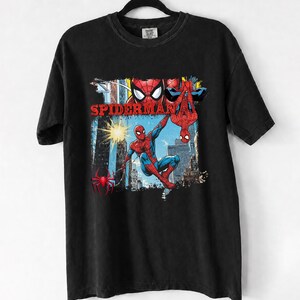 Puede incluir: Camiseta negra con un gr&aacute;fico colorido de Spider-Man en acci&oacute;n, con la palabra "SPIDERMAN" en rojo. El dise&ntilde;o incluye a Spider-Man en un paisaje urbano, con una telara&ntilde;a y una ara&ntilde;a.