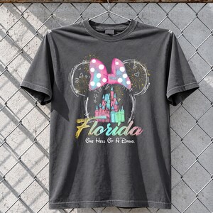 May include: Dark gray t-shirt featuring a colorful graphic of a castle, a polka-dotted bow, and the word "Florida." The text "One Hell Of A Drug" is below the word Florida. The design is in the shape of Mickey Mouse ears.