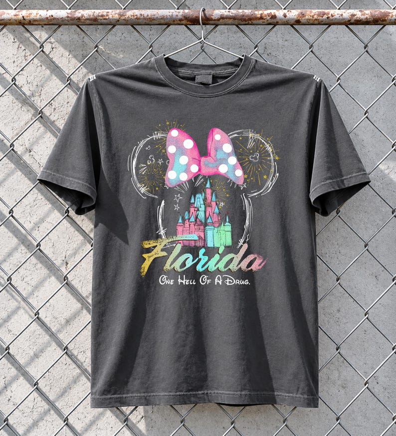 May include: Dark gray t-shirt featuring a colorful graphic of a castle, a polka-dotted bow, and the word "Florida." The text "One Hell Of A Drug" is below the word Florida. The design is in the shape of Mickey Mouse ears.