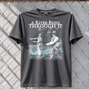 May include: Dark gray t-shirt featuring the text "A River Runs Through It" above a black and white image of two men fly fishing. The image has a vintage, faded look, and the shirt is hanging on a metal hanger.
