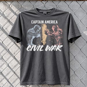 May include: Dark gray t-shirt featuring a graphic from Captain America: Civil War. The design shows two figures in action, with the words "CAPTAIN AMERICA" above and "CIVIL WAR" below in white text. The shirt is hanging on a metal hanger.