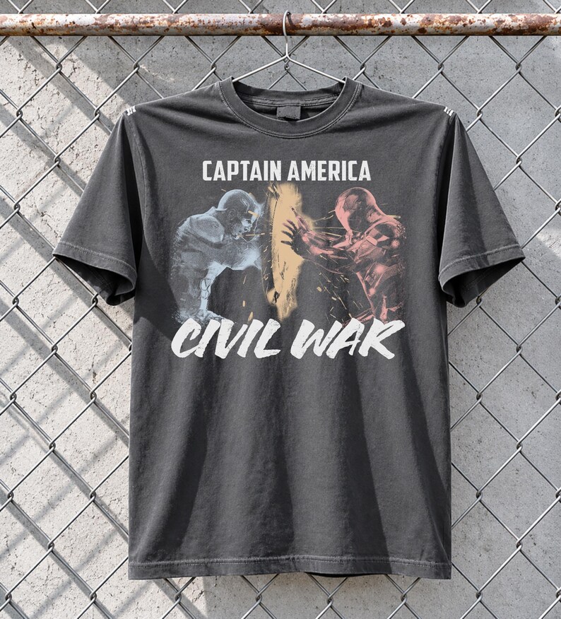 May include: Dark gray t-shirt featuring a graphic from Captain America: Civil War. The design shows two figures in action, with the words "CAPTAIN AMERICA" above and "CIVIL WAR" below in white text. The shirt is hanging on a metal hanger.