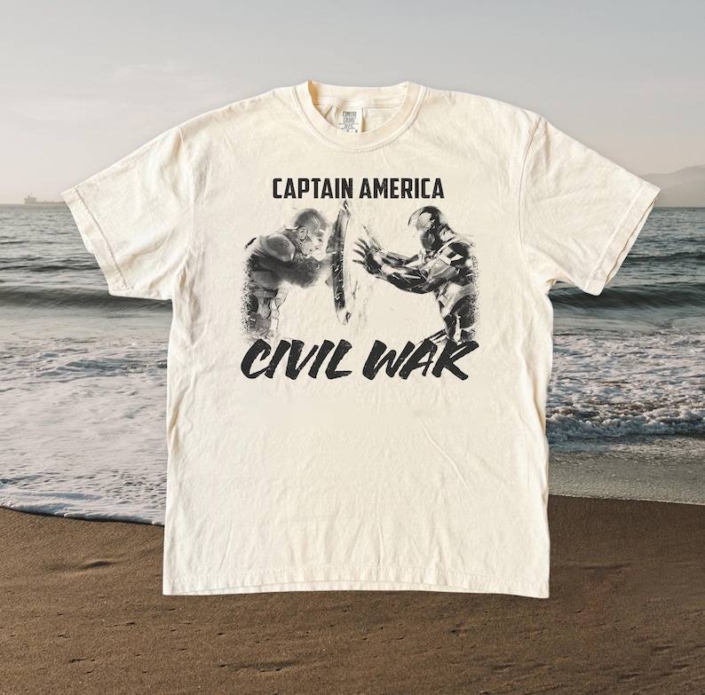 May include: Cream-colored t-shirt featuring a black and white graphic of Captain America and Iron Man in a battle pose. The text "CAPTAIN AMERICA" is above the image, and "CIVIL WAR" is below.