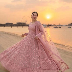 May include: A floor-length, long-sleeved dusty pink gown featuring intricate floral embroidery throughout the fabric. The dress has a full, flared skirt and is paired with a matching sheer, embroidered dupatta draped over the shoulder.