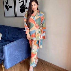 May include: A two-piece set consisting of a long-sleeved collared shirt with a tie-waist belt and matching trousers, both featuring an abstract multicoloured pattern in shades of orange, green, and blue, with the text PINK WARDROBE visible.