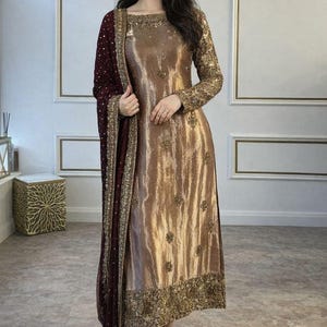 May include: A long-sleeved metallic gold tunic dress featuring scattered floral embroidery and a wide, heavily embellished gold border at the hem. The outfit includes a deep burgundy shawl with gold sequin accents and matching gold trousers.