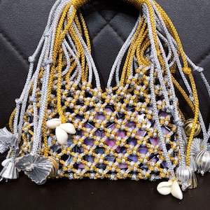Op de afbeelding: Een handgemaakte macramé tas met een goud-zilveren geweven patroon. De tas heeft een gouden handvat en bungelende zilveren en gouden koorden. Zeeschelpen en zilveren kwastjes versieren dit unieke accessoire.