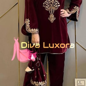 May include: Burgundy velvet two-piece outfit with gold embroidery. The long-sleeved top features ornate gold detailing around the neckline and cuffs. Matching pants and a small handbag complete the ensemble. The text "Diva Luxora" is visible.