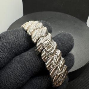 May include: Silver bracelet with a textured, woven design. The bracelet features a series of interconnected links, each with a detailed pattern. The clasp is visible, and the bracelet is displayed against a dark background.
