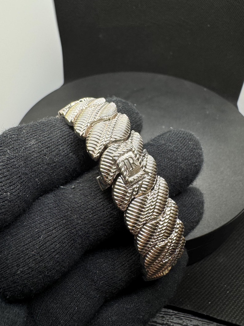 May include: Silver bracelet with a textured, woven design. The bracelet features a series of interconnected links, each with a detailed pattern. The clasp is visible, and the bracelet is displayed against a dark background.