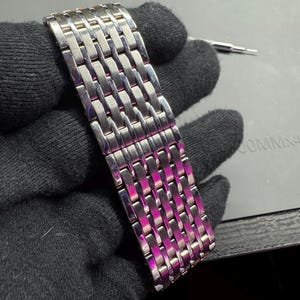 May include: A silver-toned watch band with a woven design. The band is made of rectangular links that are interlocked. The band is held by a gloved hand. A small tool is visible in the background.