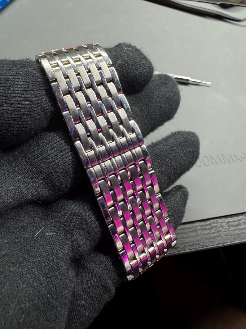 May include: A silver-toned watch band with a woven design. The band is made of rectangular links that are interlocked. The band is held by a gloved hand. A small tool is visible in the background.